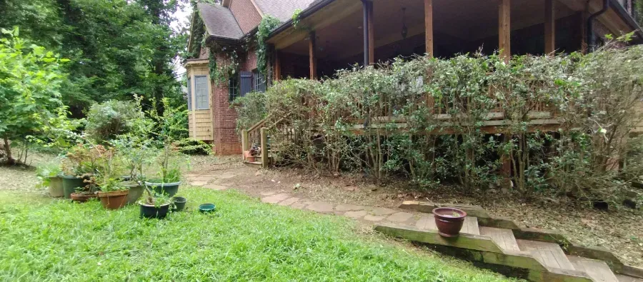 A brick house with a wooden porch sits behind a line of hedges, featuring a garden with potted plants and stone steps.