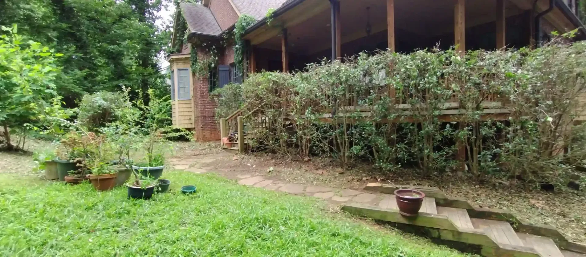 A rustic house with a wooden porch is partially hidden behind a long hedge, alongside stone steps leading to a green lawn.