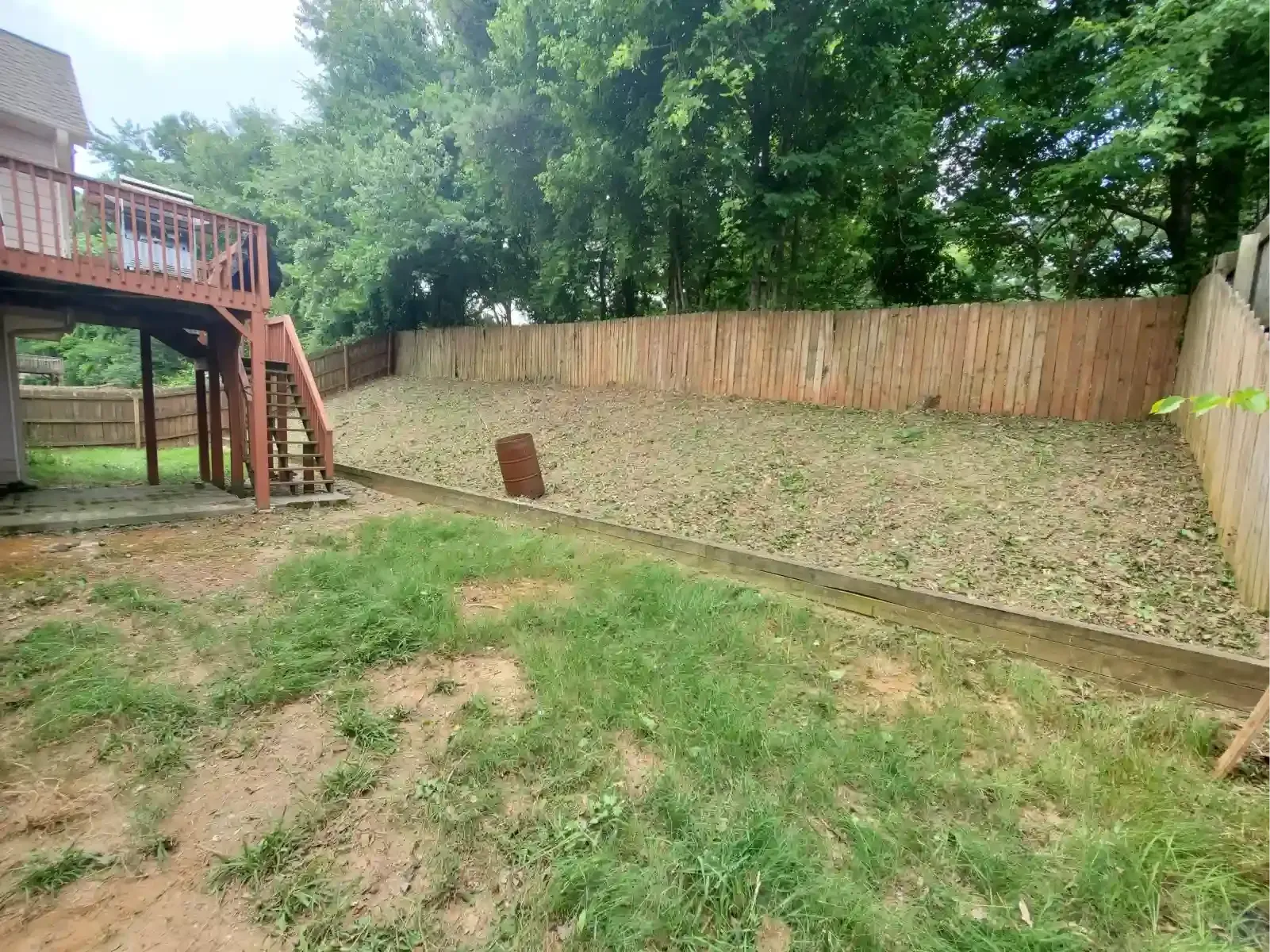 A sloped backyard with a wooden fence, a grass and dirt lawn, and a multi-story wooden deck extending from a house.