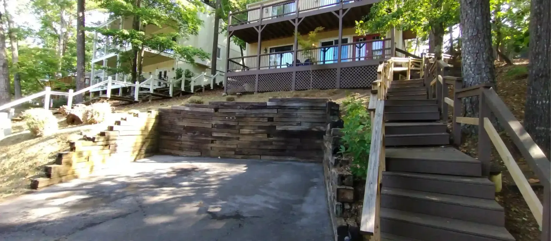 A paved parking area in front of a wooden retaining wall and a set of stairs leading to a lakefront house.