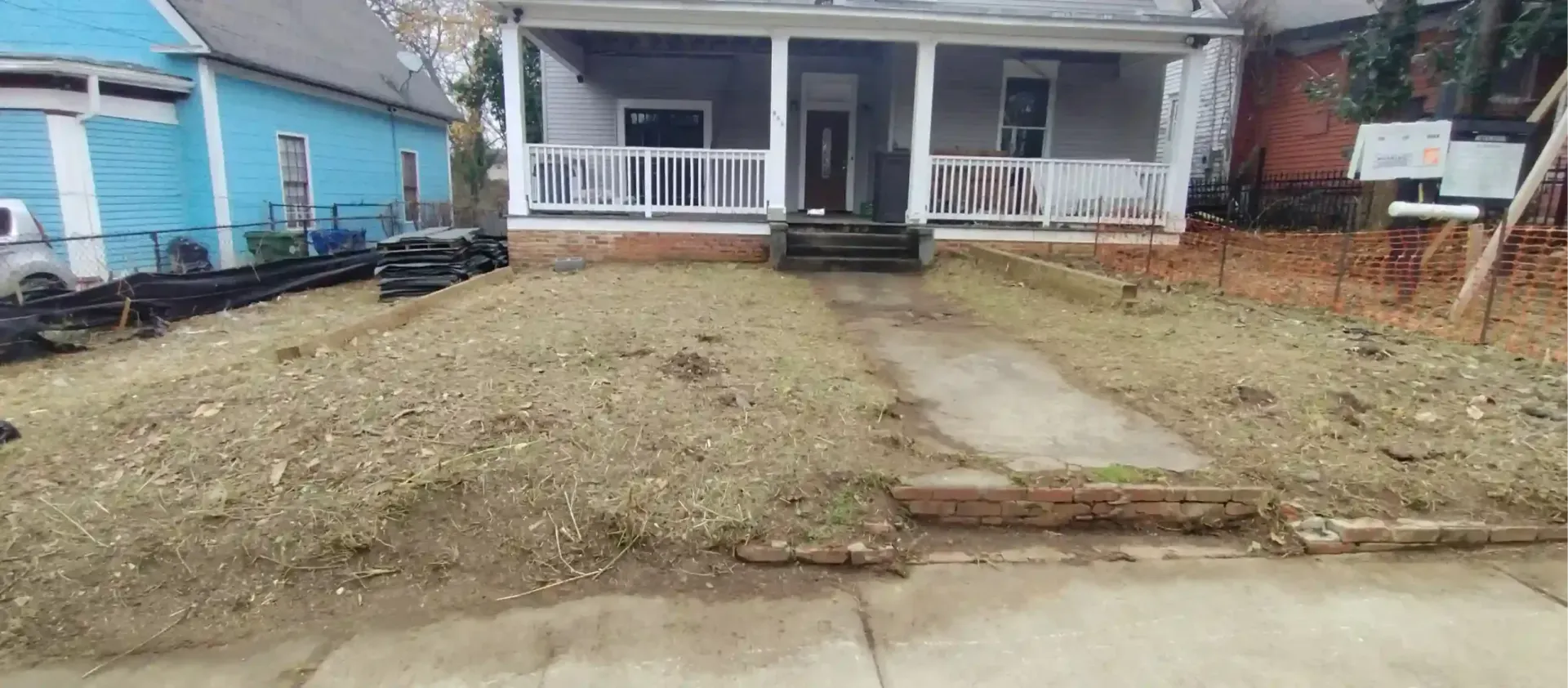 A gray house with a front porch, white railing, and stairs, viewed from the street with a patch of dirt front lawn.