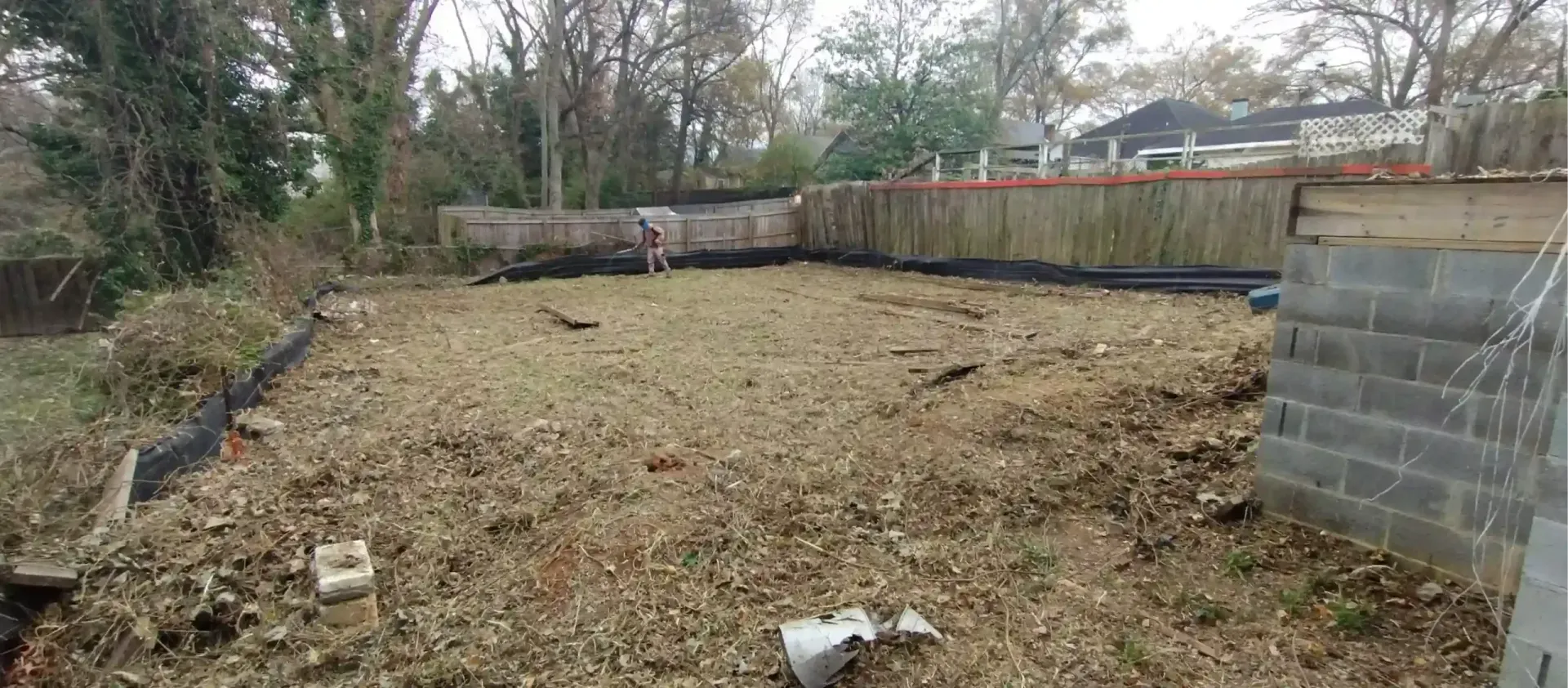 A cleared, uneven dirt lot with scattered leaves, enclosed by a wooden fence and a partially built concrete block wall.