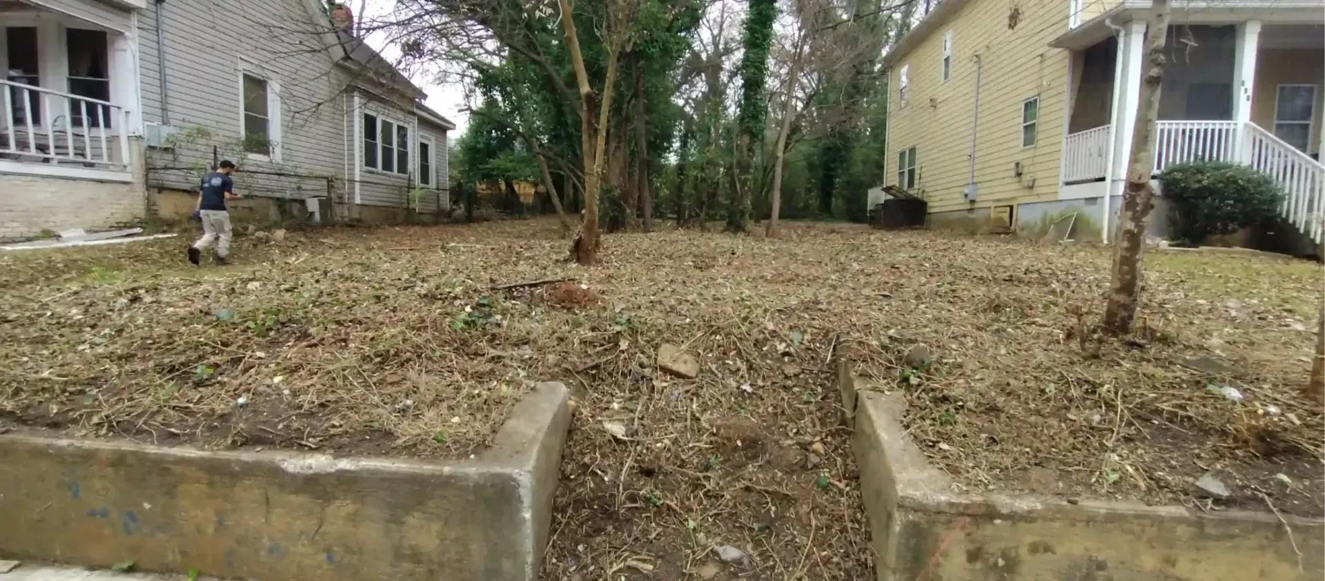 A leaf-covered yard between two houses, featuring a concrete retaining wall with a central opening at the front.
