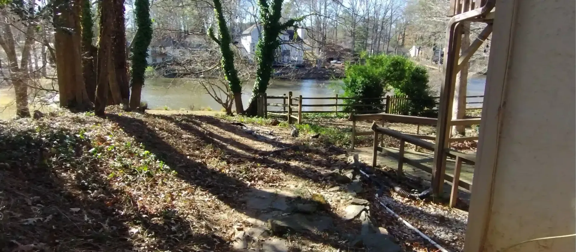 A sunlit, wooded yard with fallen leaves, a rustic wooden fence, and a calm river in the background.