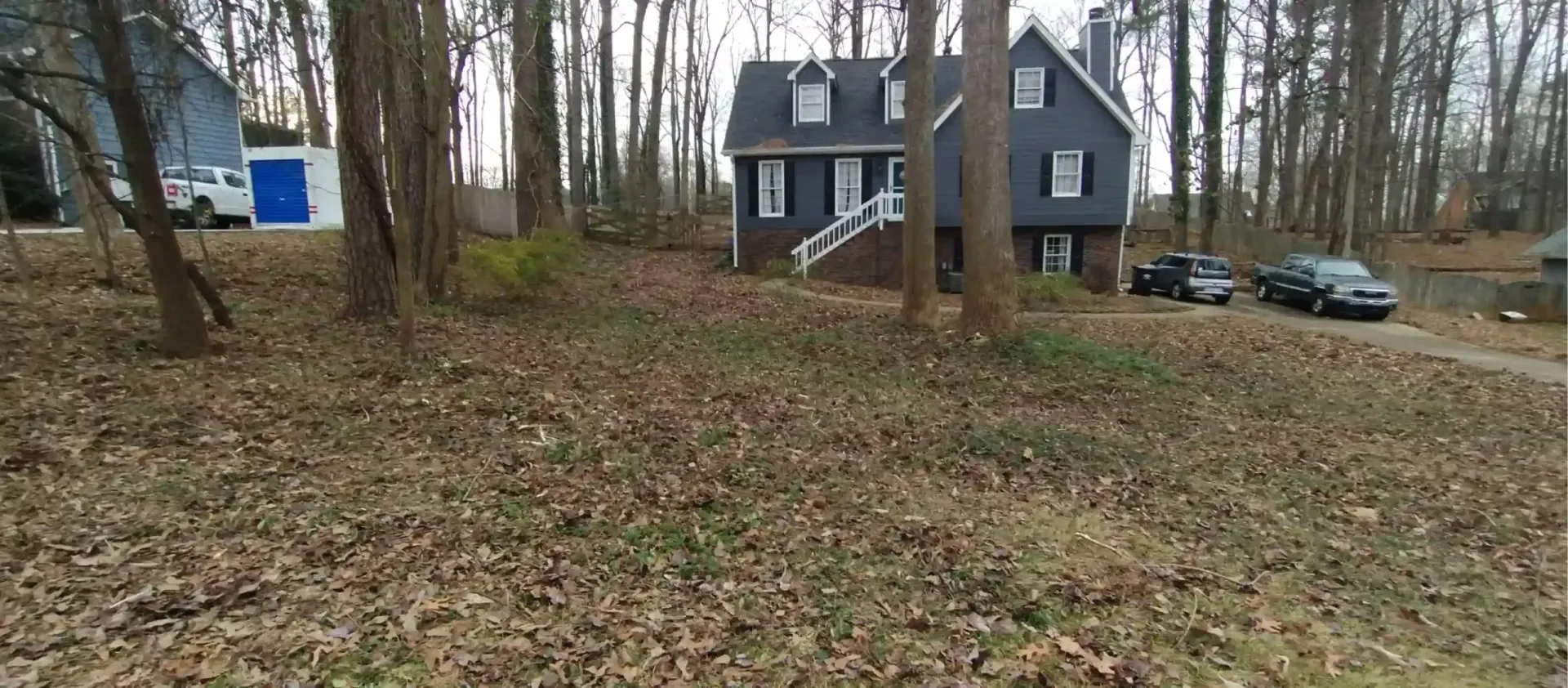A grey two-story house with a white staircase, surrounded by trees and leaf-covered ground, with cars parked nearby.
