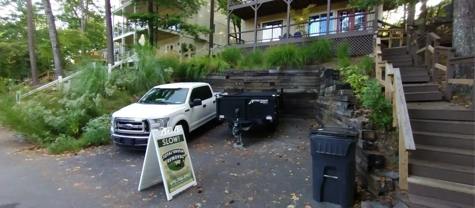 A white pickup truck parked in a driveway next to a small utility trailer and a sign, with stairs leading to a house.
