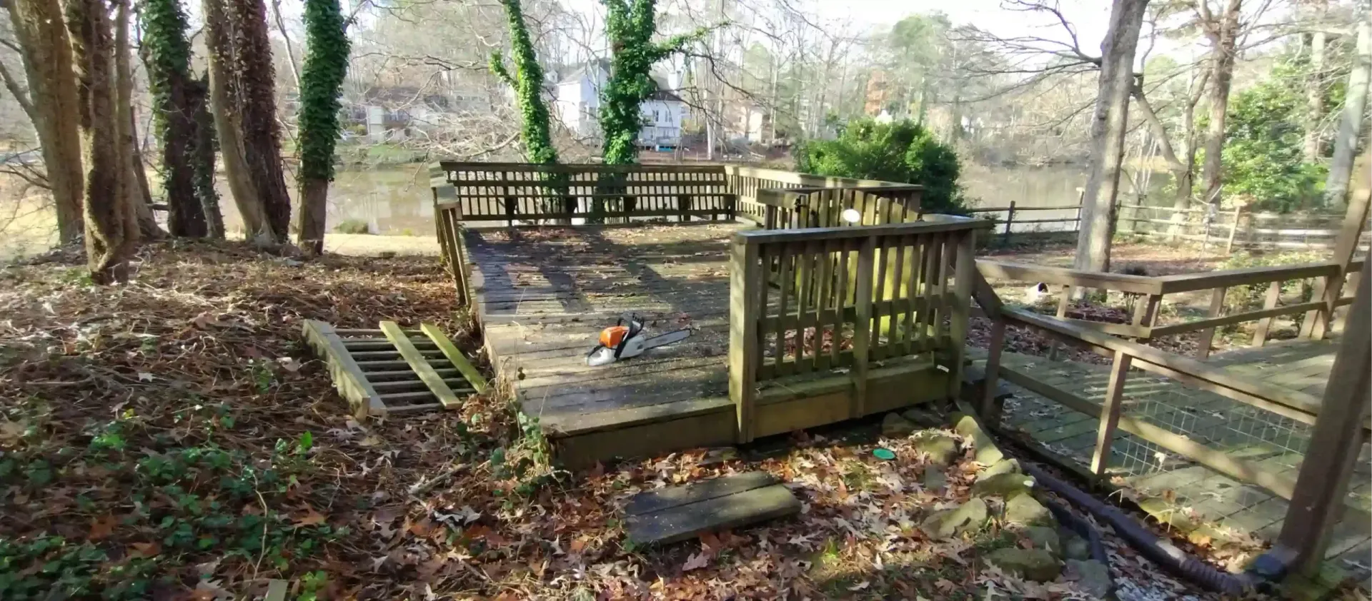A wooden deck with railings overlooks a pond surrounded by trees on a sunny day with fallen leaves on the ground.