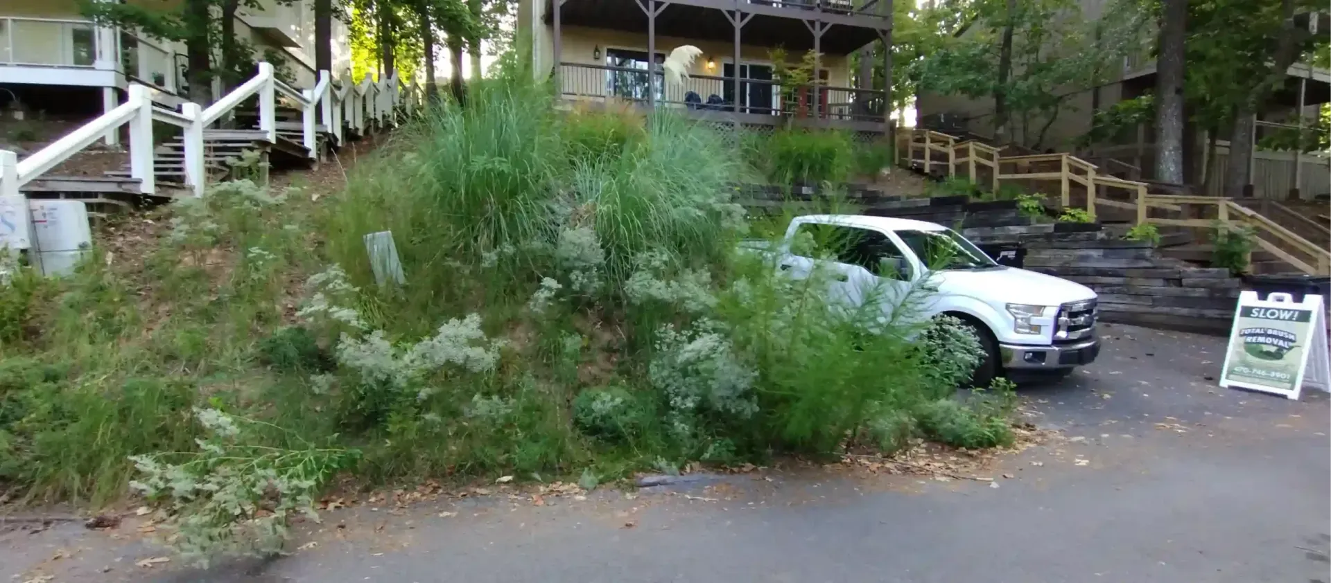 A white pickup truck parked next to a grassy, sloped hill with wooden fences and stairs in a residential setting.