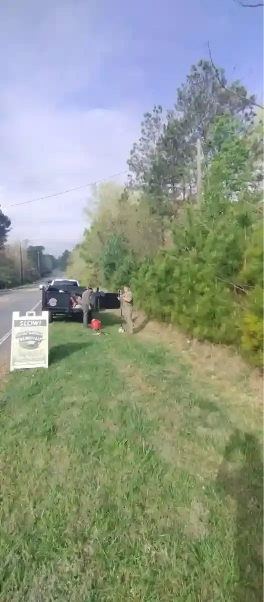 A dark SUV is parked on a grassy shoulder next to a sign for a wood-cutting service, with two people working nearby.
