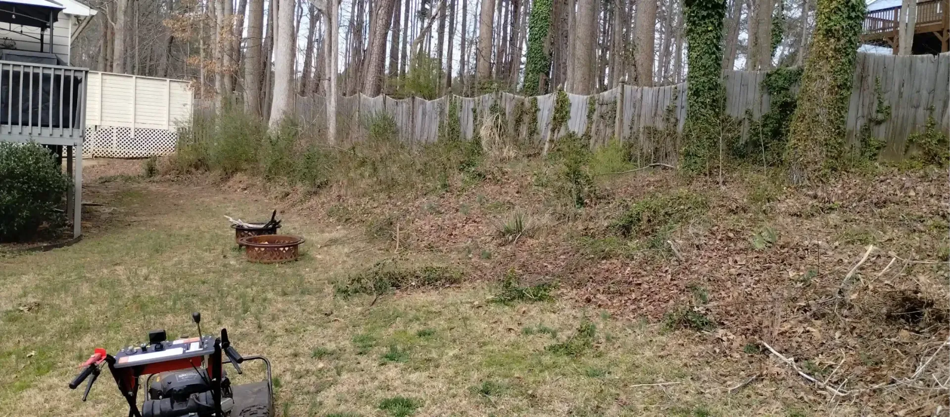 A lawnmower sits on a grassy yard next to a sloped area covered in leaves and brush near a wooded fence.