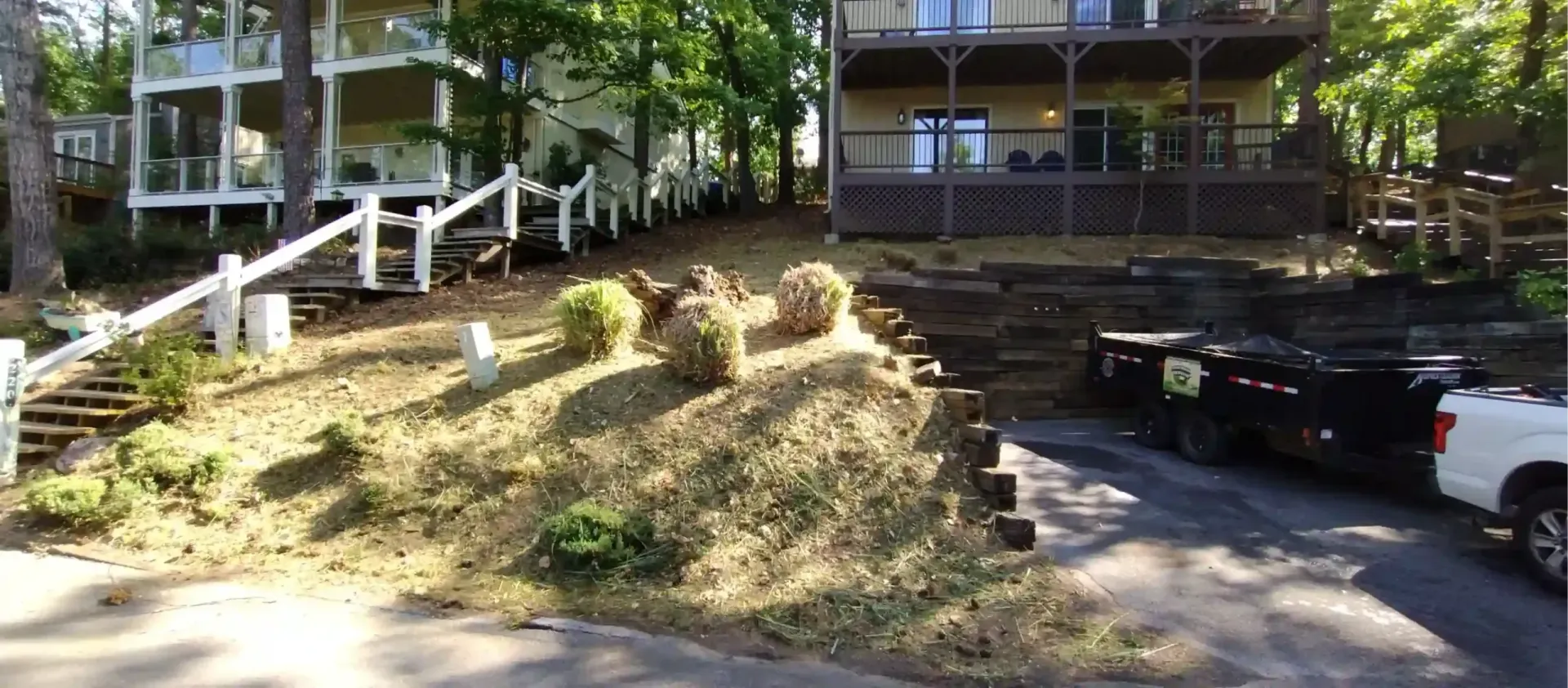 A sloped, landscaped residential yard with stairs, a stone retaining wall, and a truck parked on a driveway.