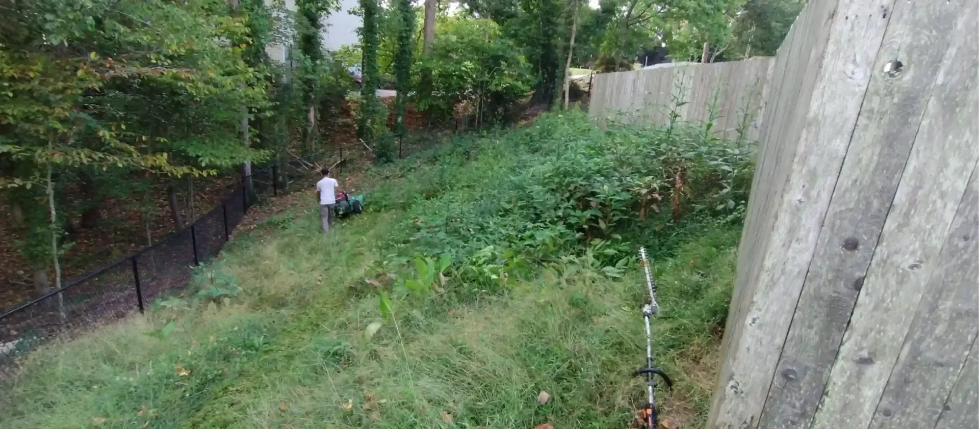 A person uses a string trimmer to clear overgrown grass and weeds on a sloped hillside next to a concrete retaining wall.