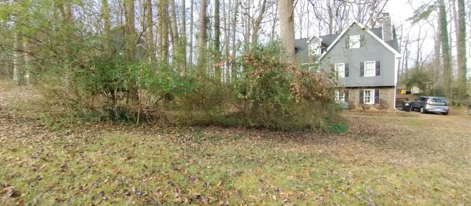 A gray, multi-story house sits in a wooded yard with fallen branches and dried leaves covering the grass.
