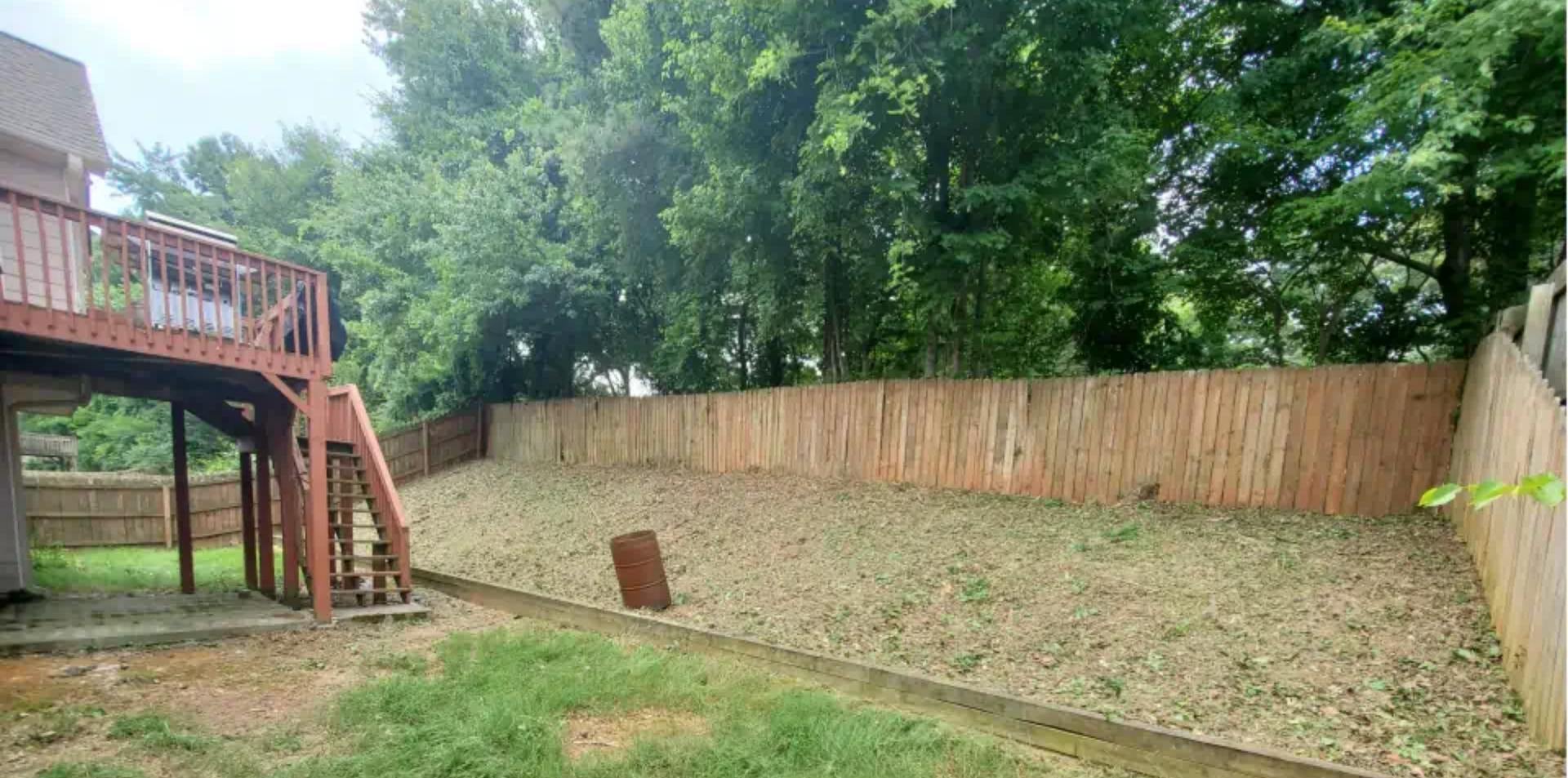A sloped backyard featuring a wooden deck with stairs on the left, a tall wooden privacy fence, and scattered foliage.