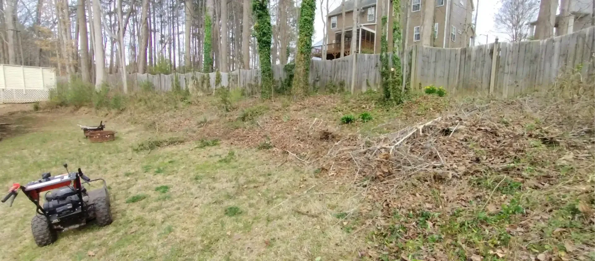 A walk-behind brush mower sits on a lawn near a sloped, wooded area with a house and fence in the background.
