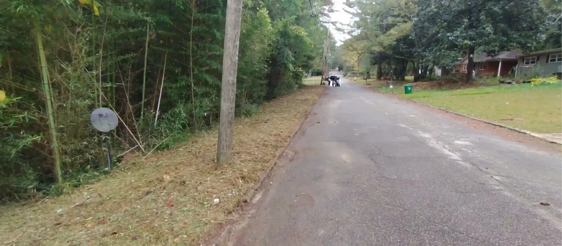 A paved road stretches into a wooded neighborhood with grass, trees, a satellite dish, and figures walking in the distance.