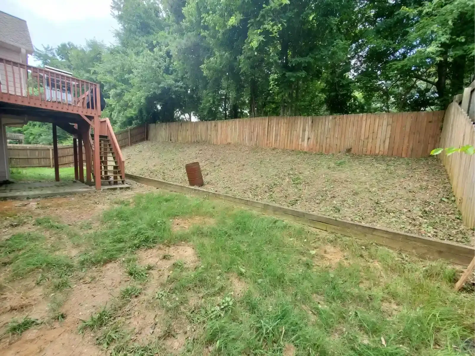 A wooden deck overlooks a grassy backyard with a sloped hill, a retaining wall, and a wooden fence.