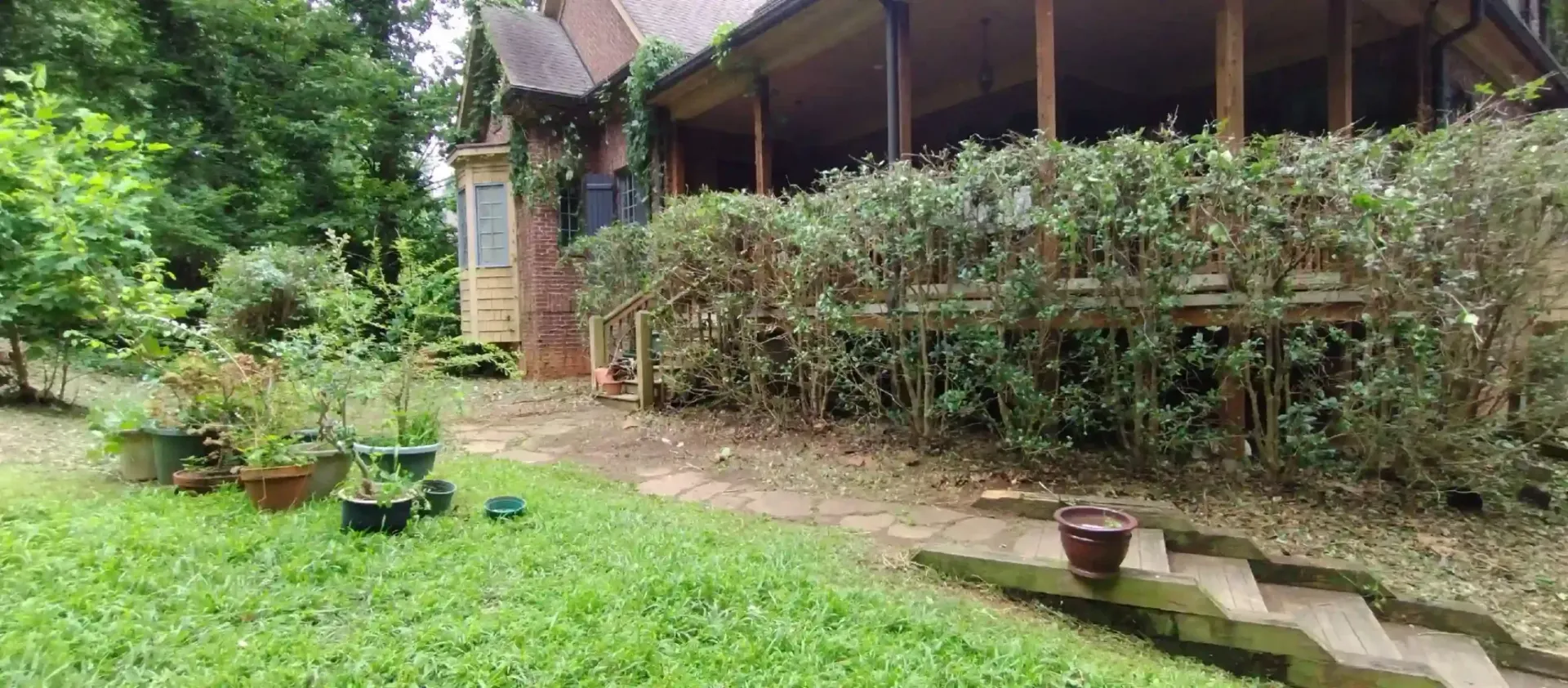 A brown house with a wrap-around porch, surrounded by lush green shrubs and trees, with stone steps in the foreground.