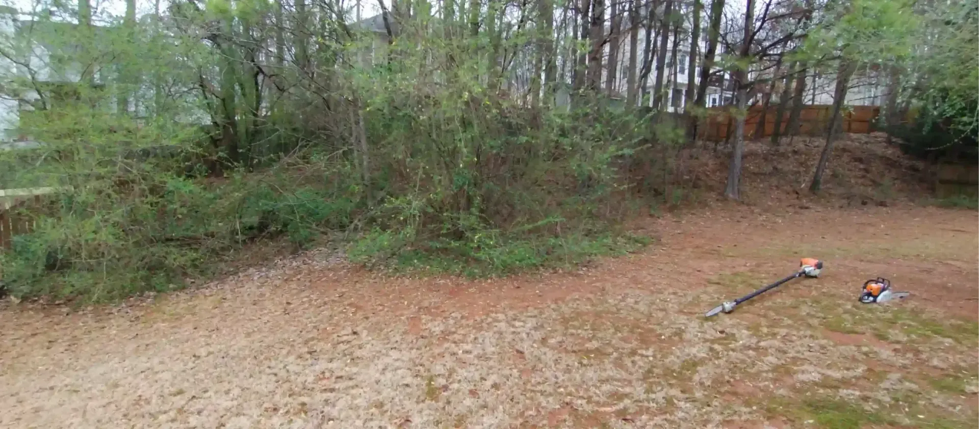 A string trimmer and its blower attachment lie on a pine-needle-covered yard in front of a wooded area.