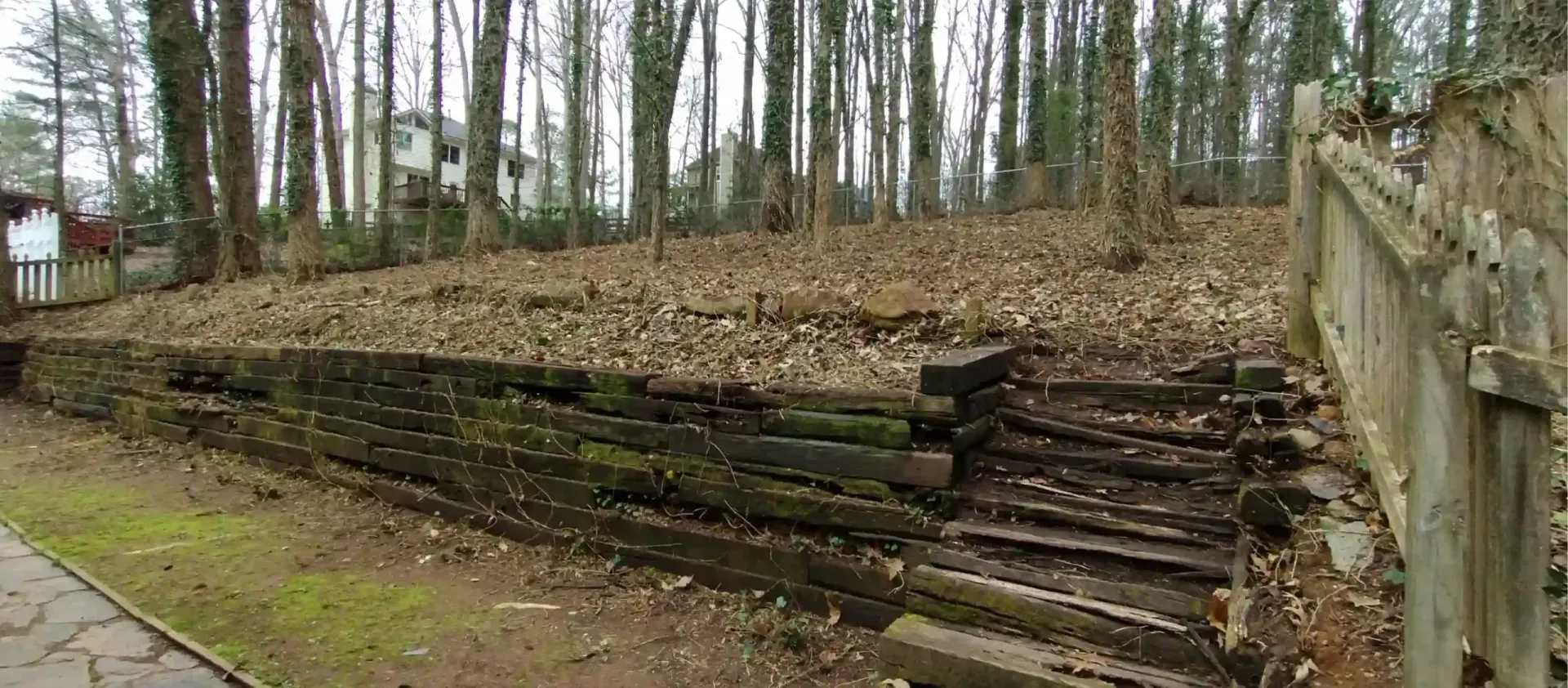 An old wooden retaining wall with integrated steps leading up a sloped, leaf-covered wooded yard.