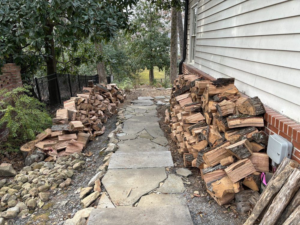 A stone pathway leads downhill between two large stacks of firewood next to the side of a house.