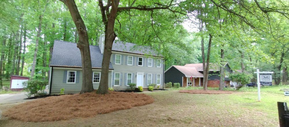 A two-story grey colonial house with a dark roof and a separate dark structure set in a lush, tree-filled yard.