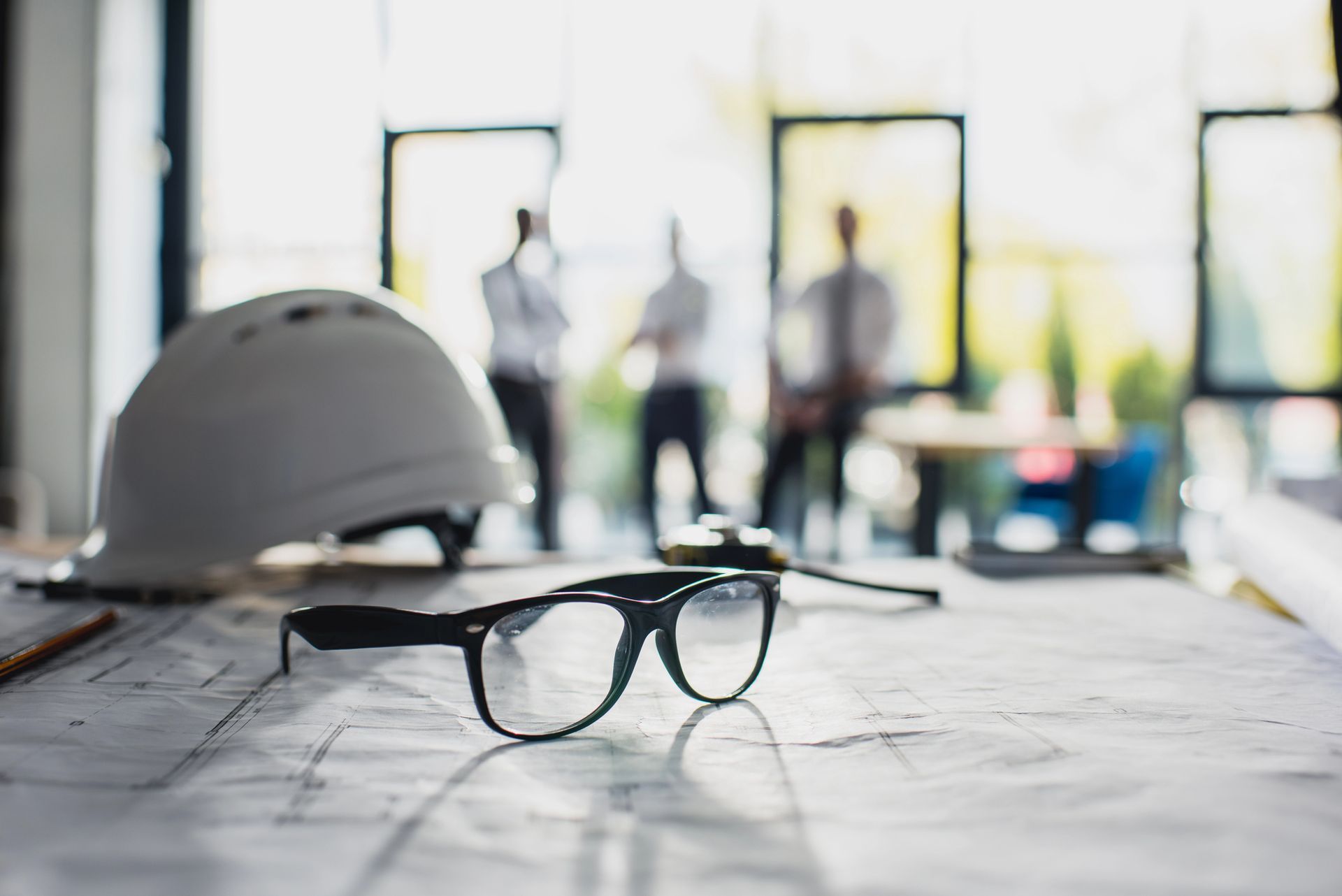 Gafas negras sobre una mesa con un casco blanco en un fondo borroso de oficina o lugar de trabajo.