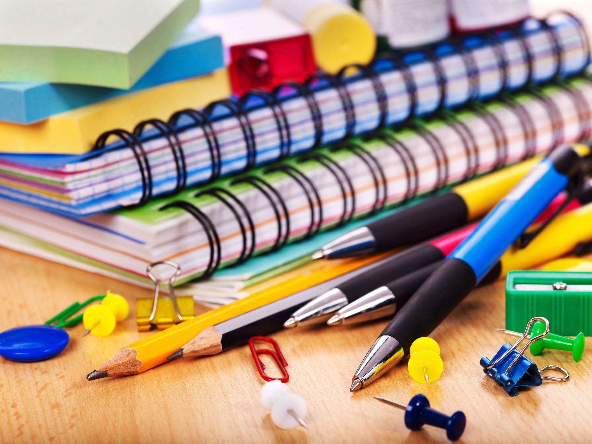 Material escolar colorido: cuadernos de espiral, lápices, bolígrafos, clips y chinchetas sobre un escritorio.
