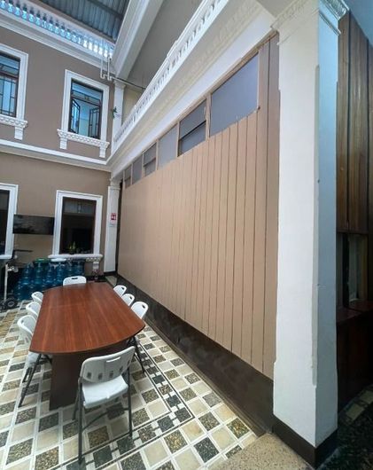 Estrecha zona de comedor interior con mesa larga de madera, sillas blancas, suelo de baldosas y paneles de pared beige.