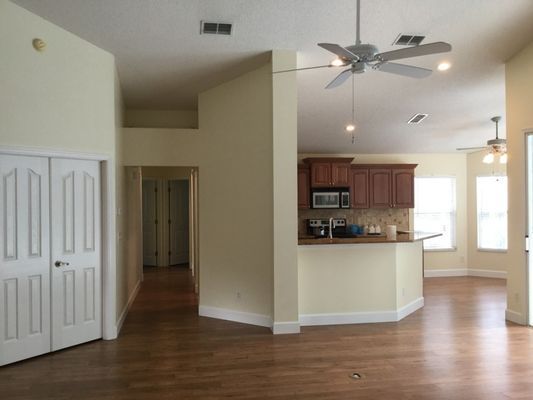 Empty living room with hardwood floors, open kitchen, ceiling fan, and hallway entrance