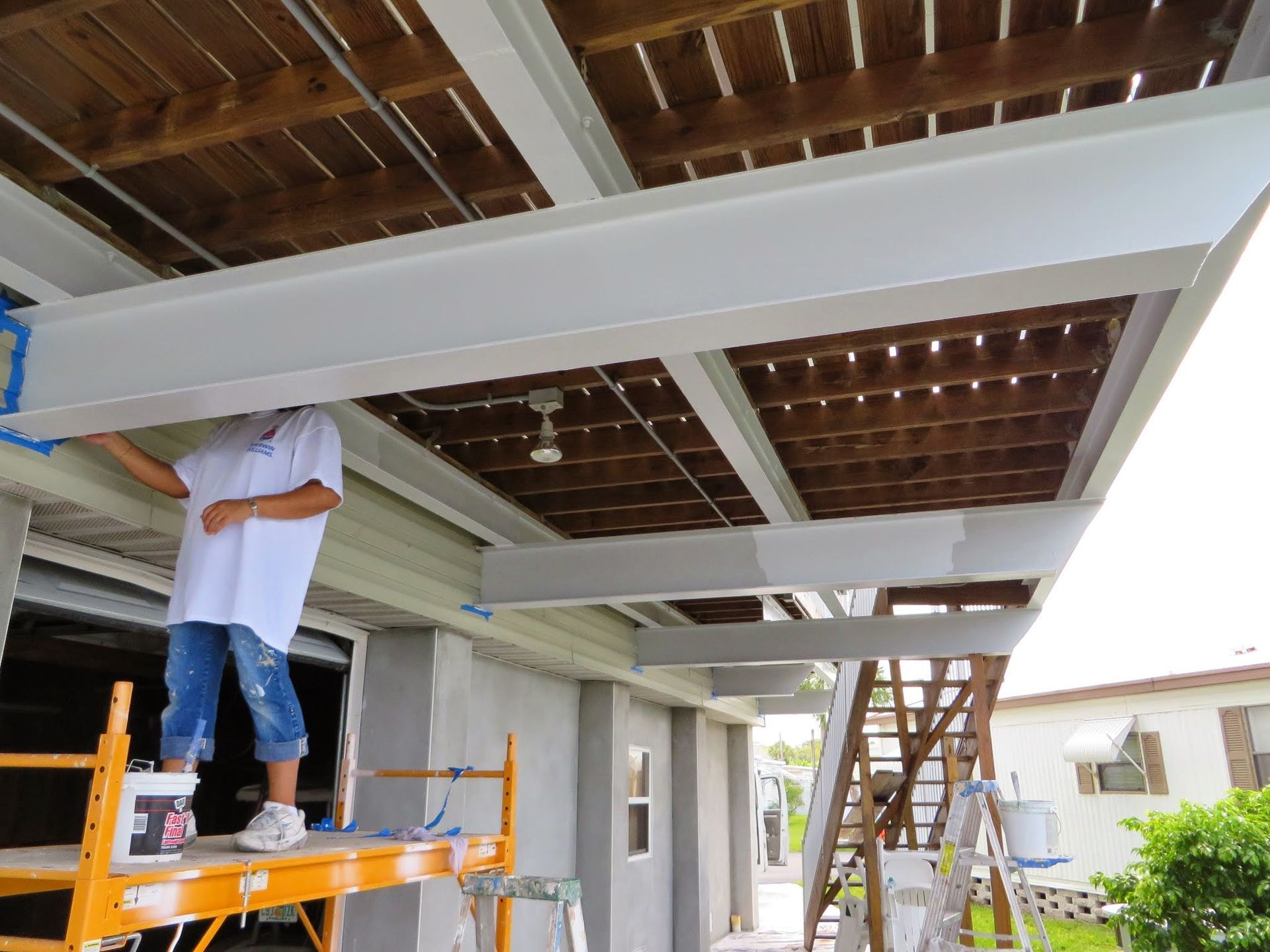 Worker on orange scaffold installing white beams under a covered patio ceiling.