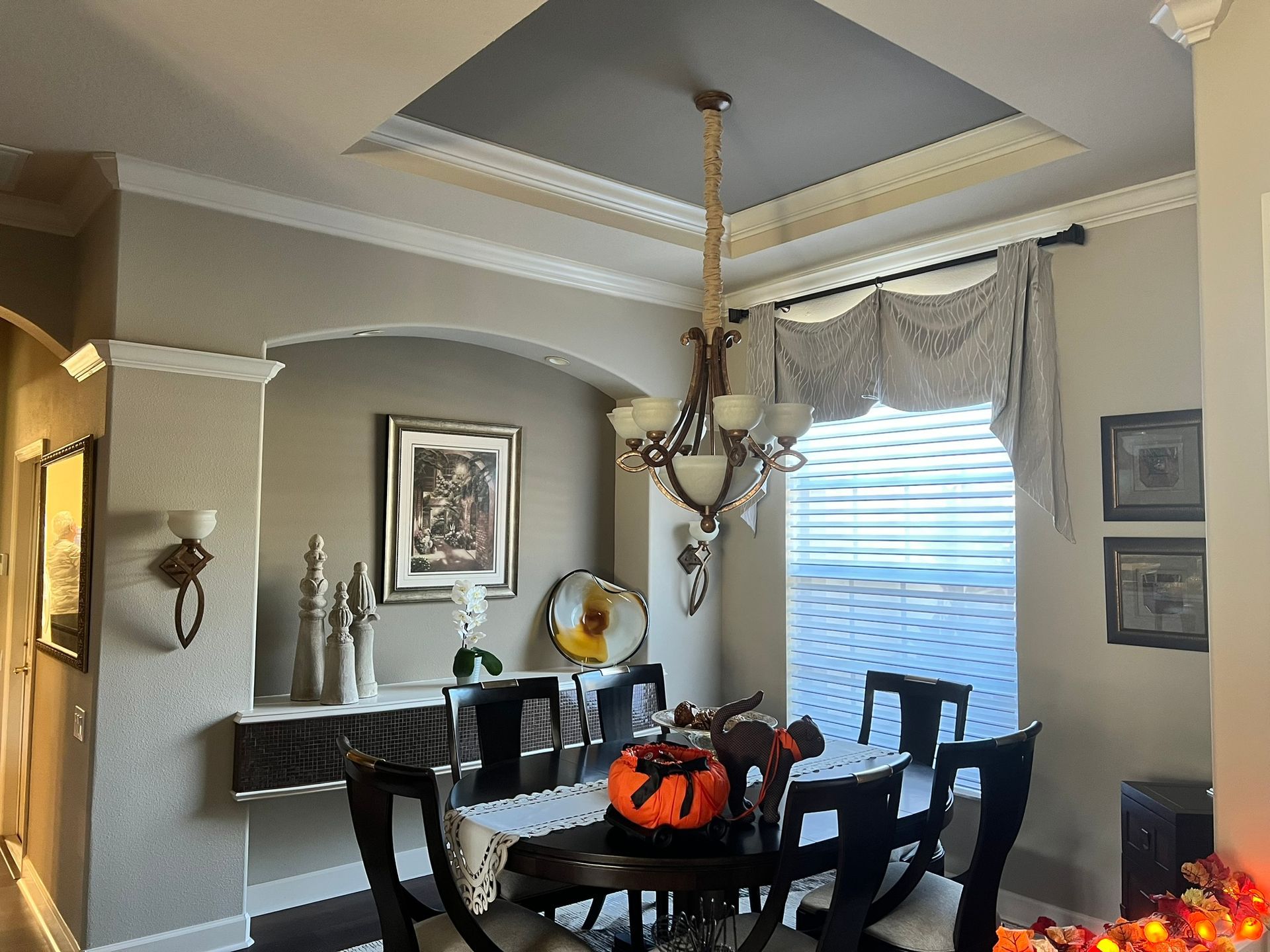 Dining room with chandelier, dark wood table, autumn centerpiece, and large window with draped curtains.