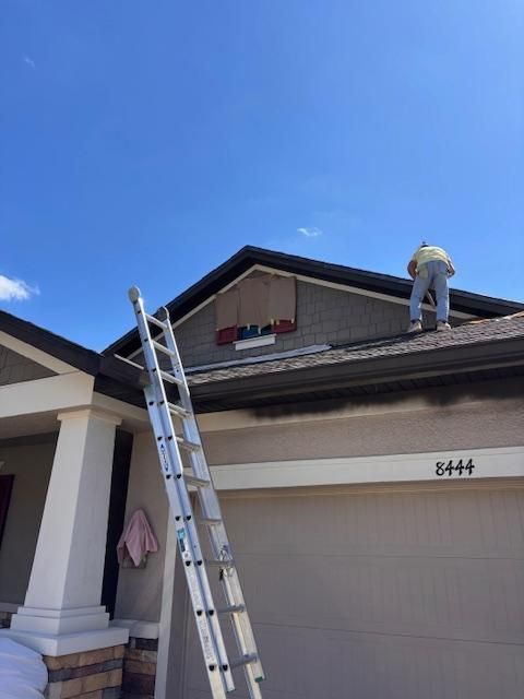 Worker on a roof beside a ladder above a suburban garage, with blue sky overhead.