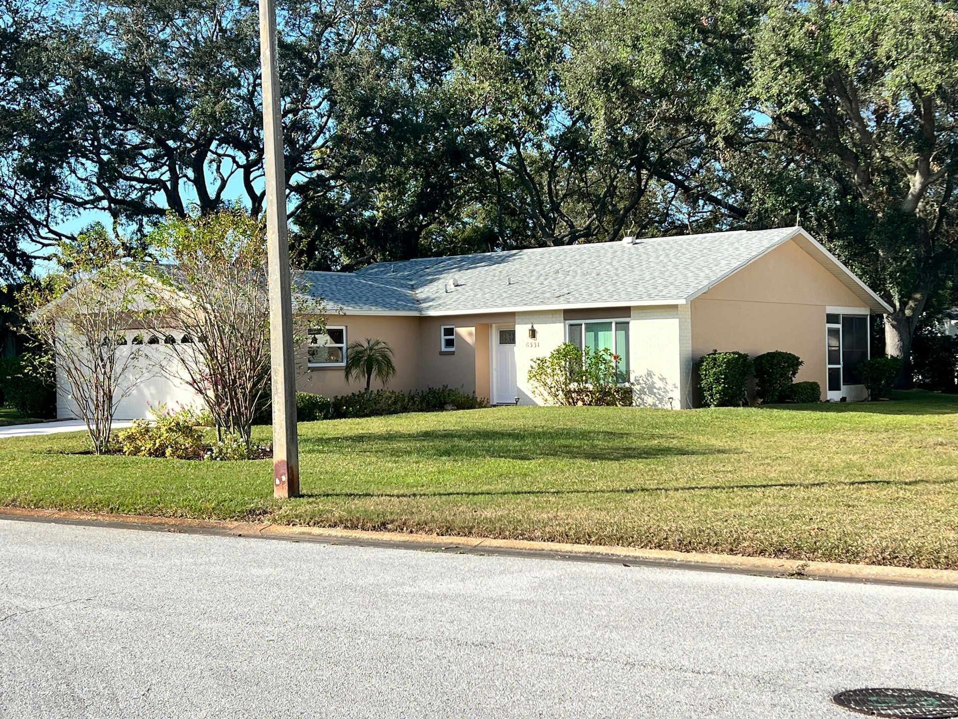 Single-story beige house with a lawn, trees, and a utility pole beside the street