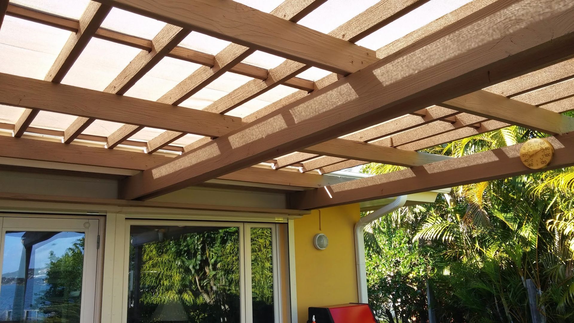 Covered patio with wooden pergola roof, yellow wall, and sliding glass doors overlooking greenery