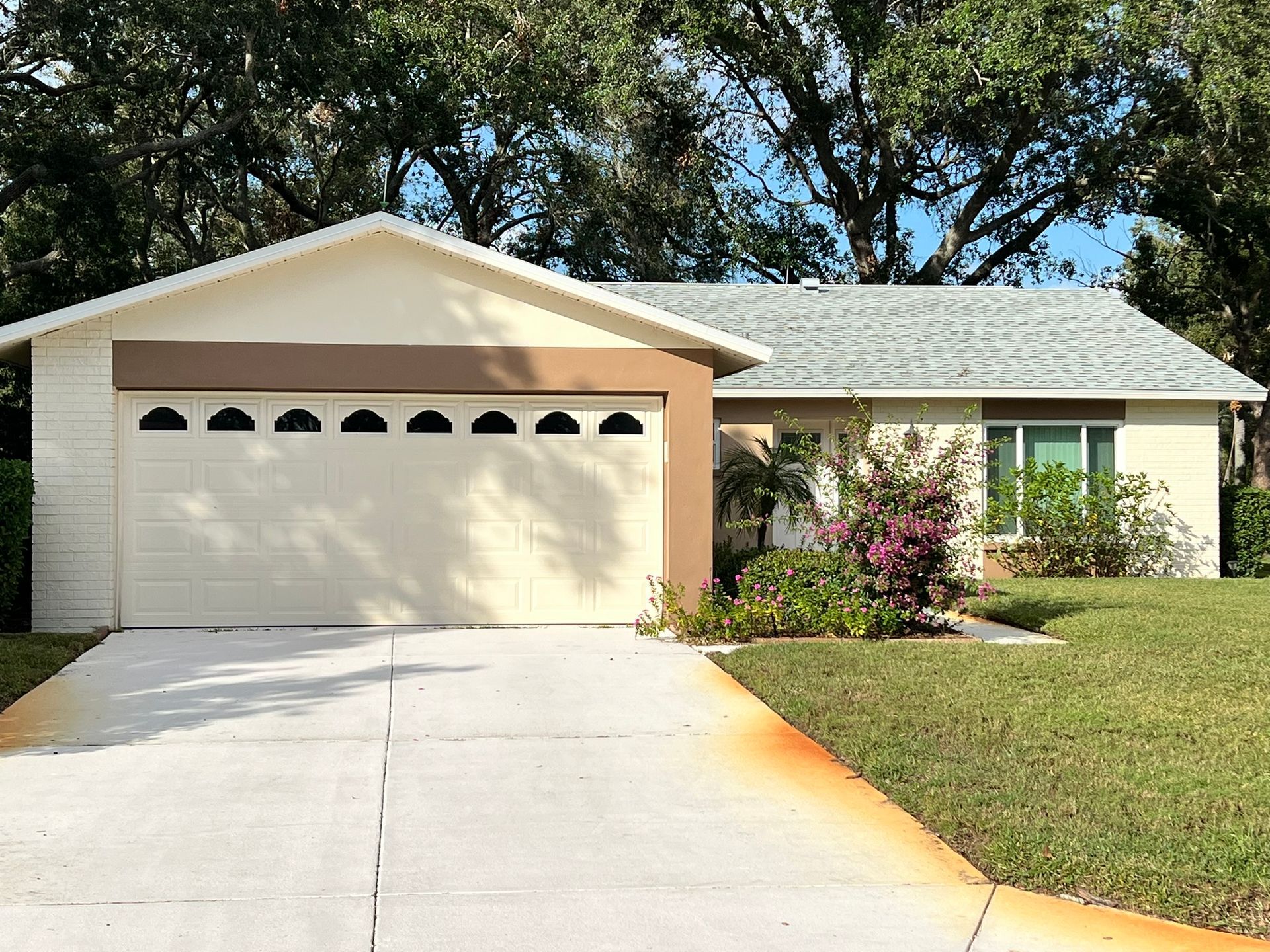 Single-story beige house with a two-car garage, driveway, and front garden under trees
