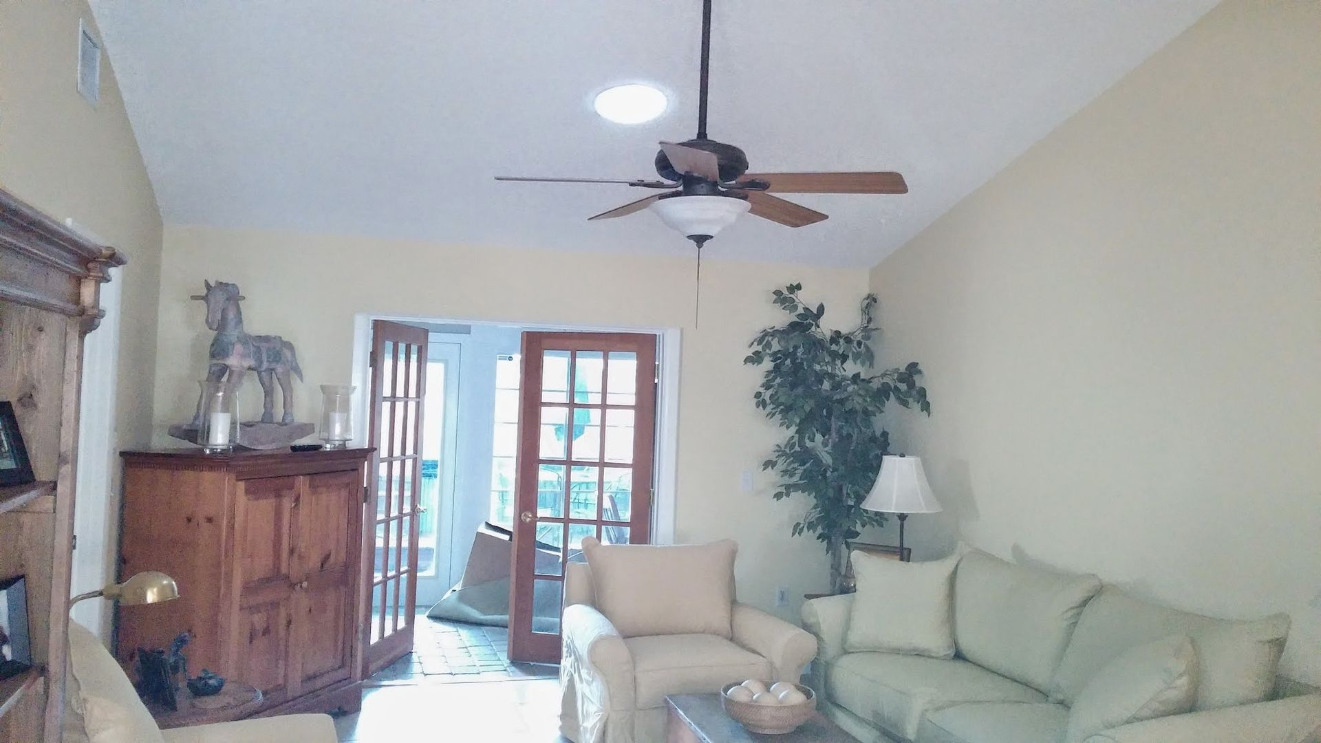 Bright living room with beige sofa, armchair, wooden cabinet, ceiling fan, and glass doors to a patio.
