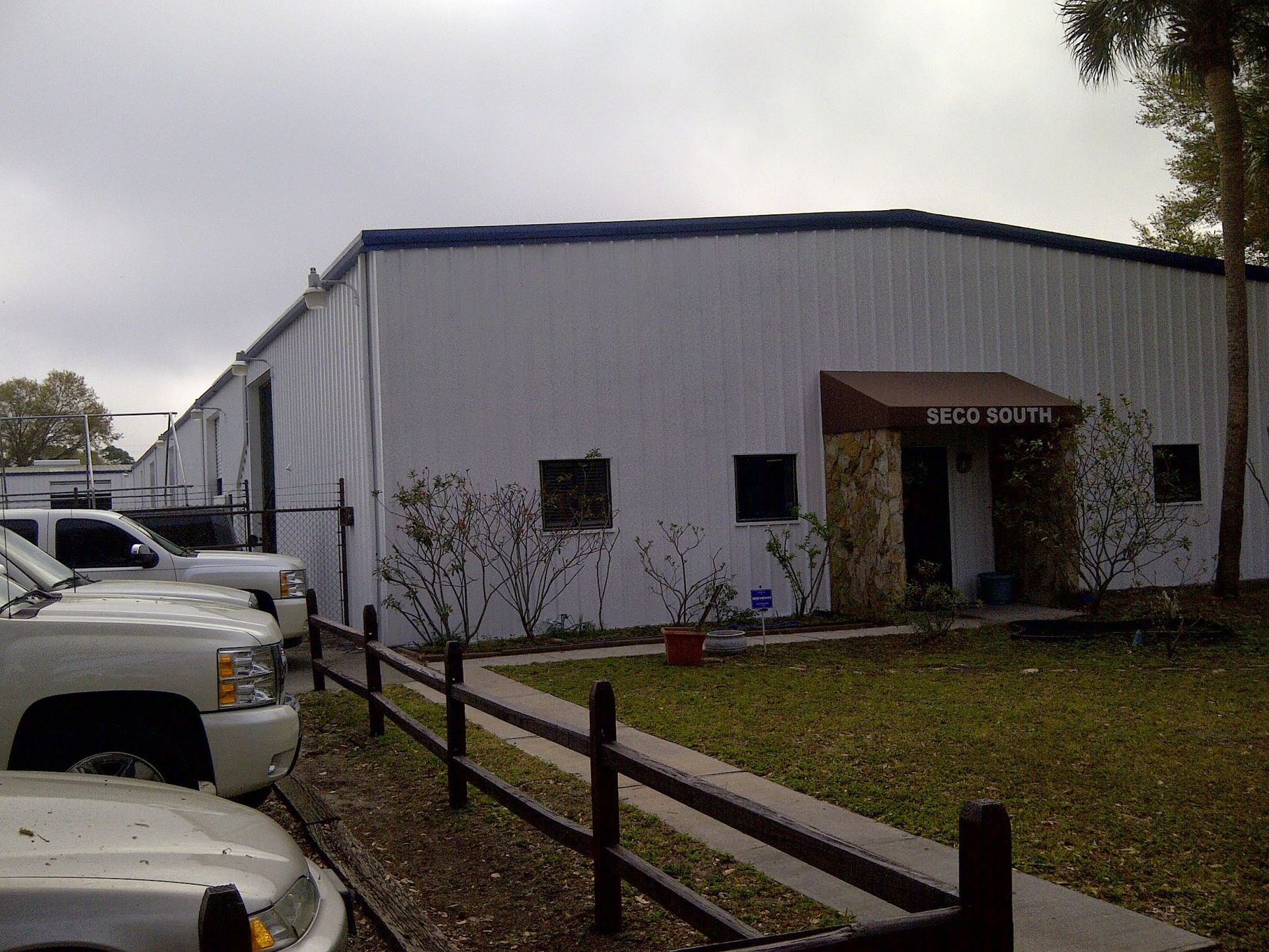 White industrial building with a small entrance, parked trucks, and a wooden fence along a gravel driveway.