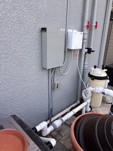 Gray utility boxes and white pipes mounted on an exterior wall near pool equipment and a hose reel.