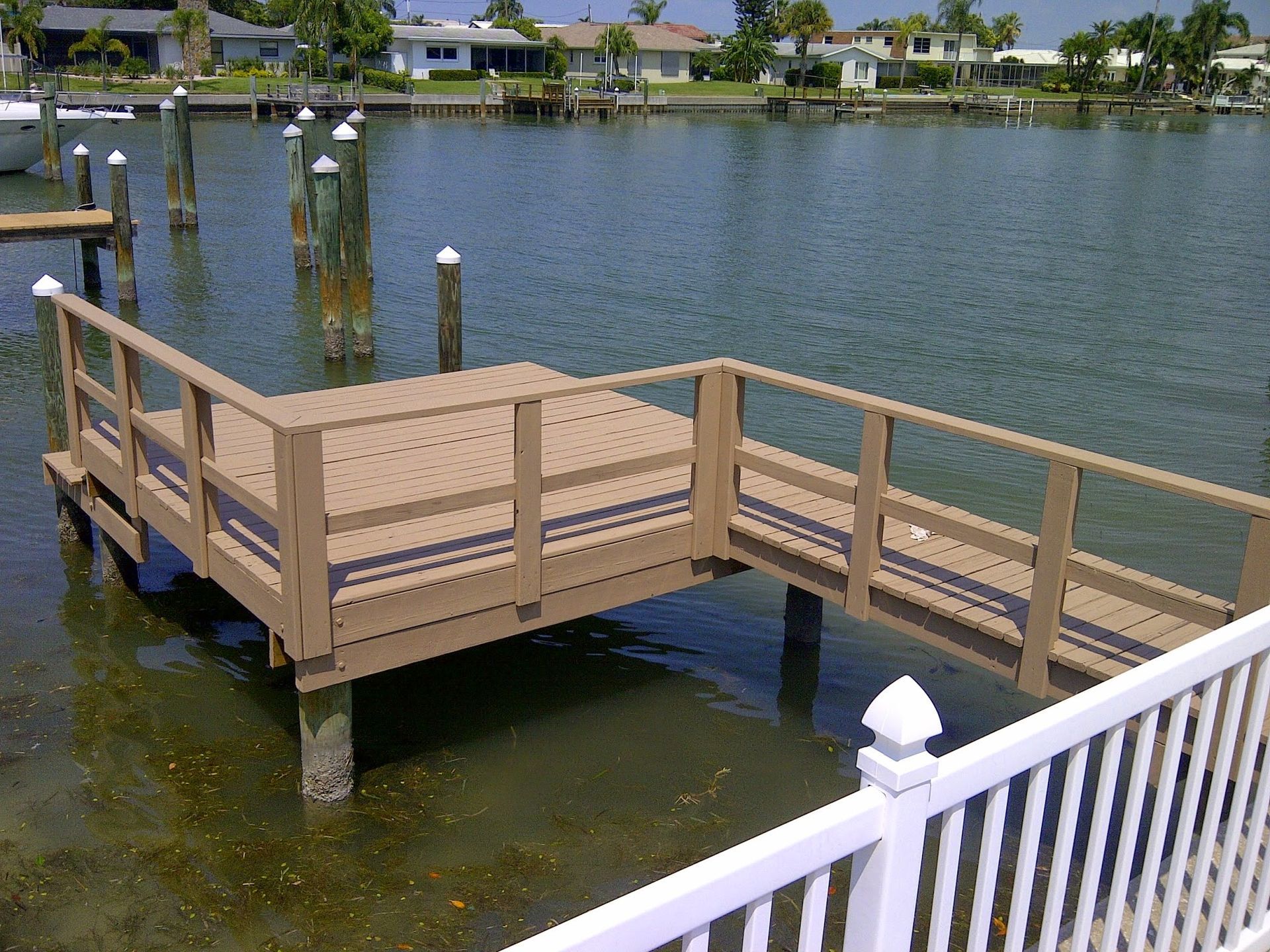 Small wooden dock with railings extending over calm water near waterfront homes.
