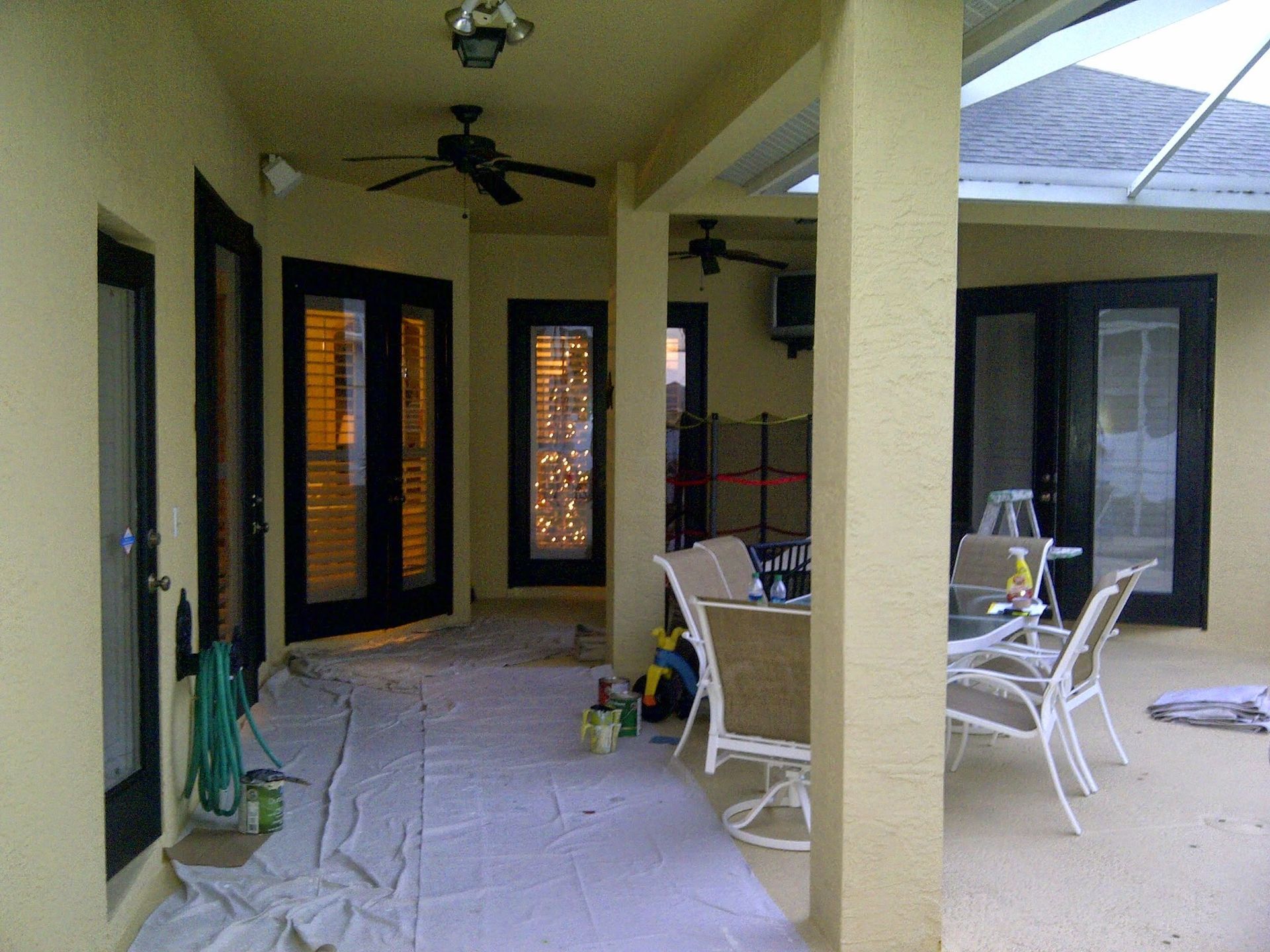 Covered patio with white chairs, pillars, and ceiling fans beside dark-framed glass doors.