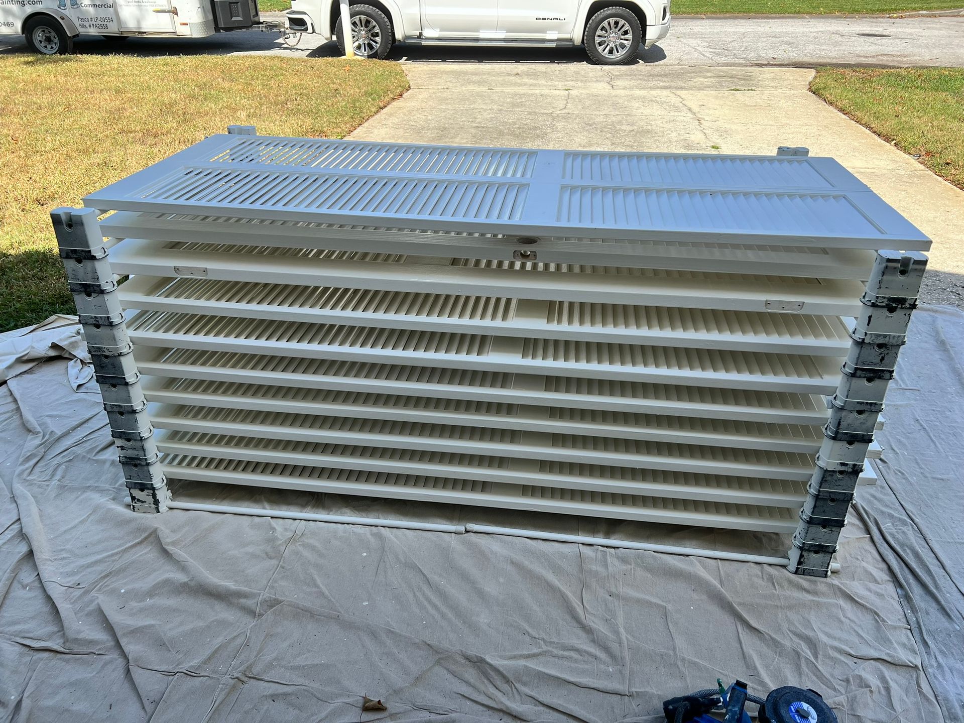 Stacked white metal panels on a tarp outdoors, with cars and a driveway in the background