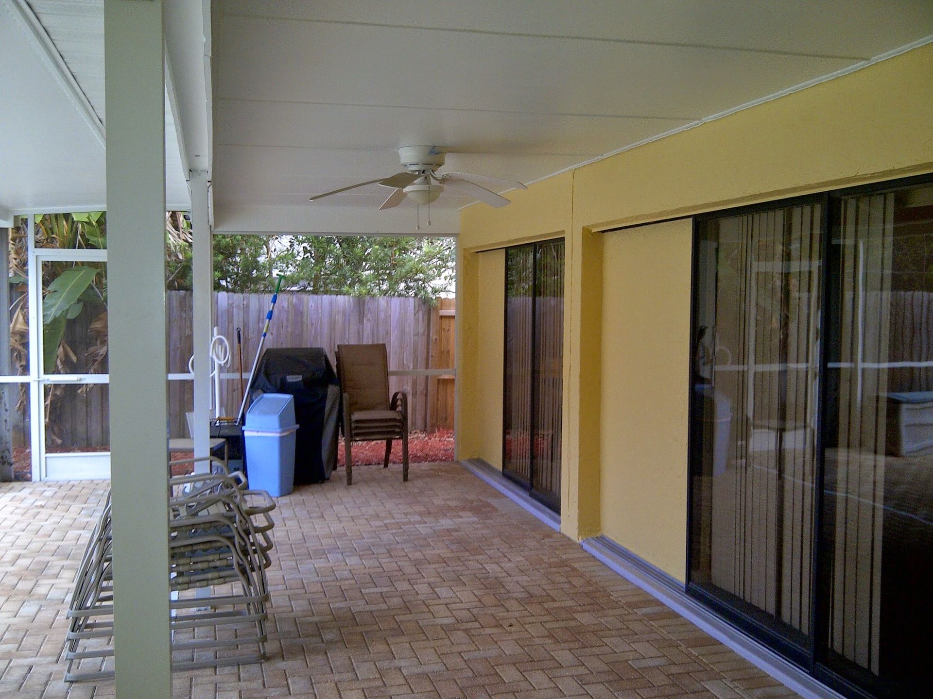 Covered patio with white ceiling, yellow wall, sliding glass doors, brick floor, chairs, grill, and blue bin