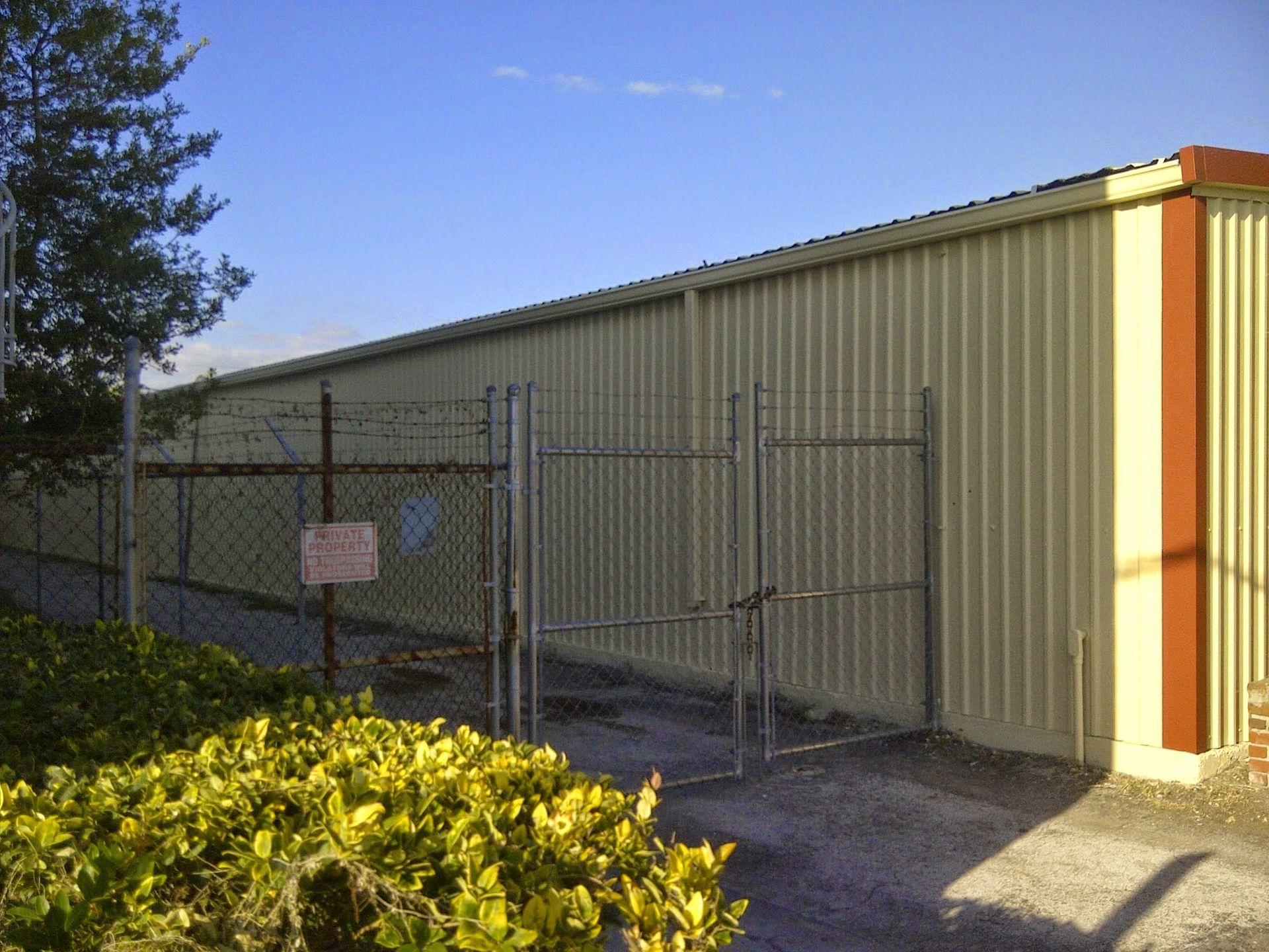 Chain-link fence in front of a tan metal building with a gate and shrubs in the foreground.