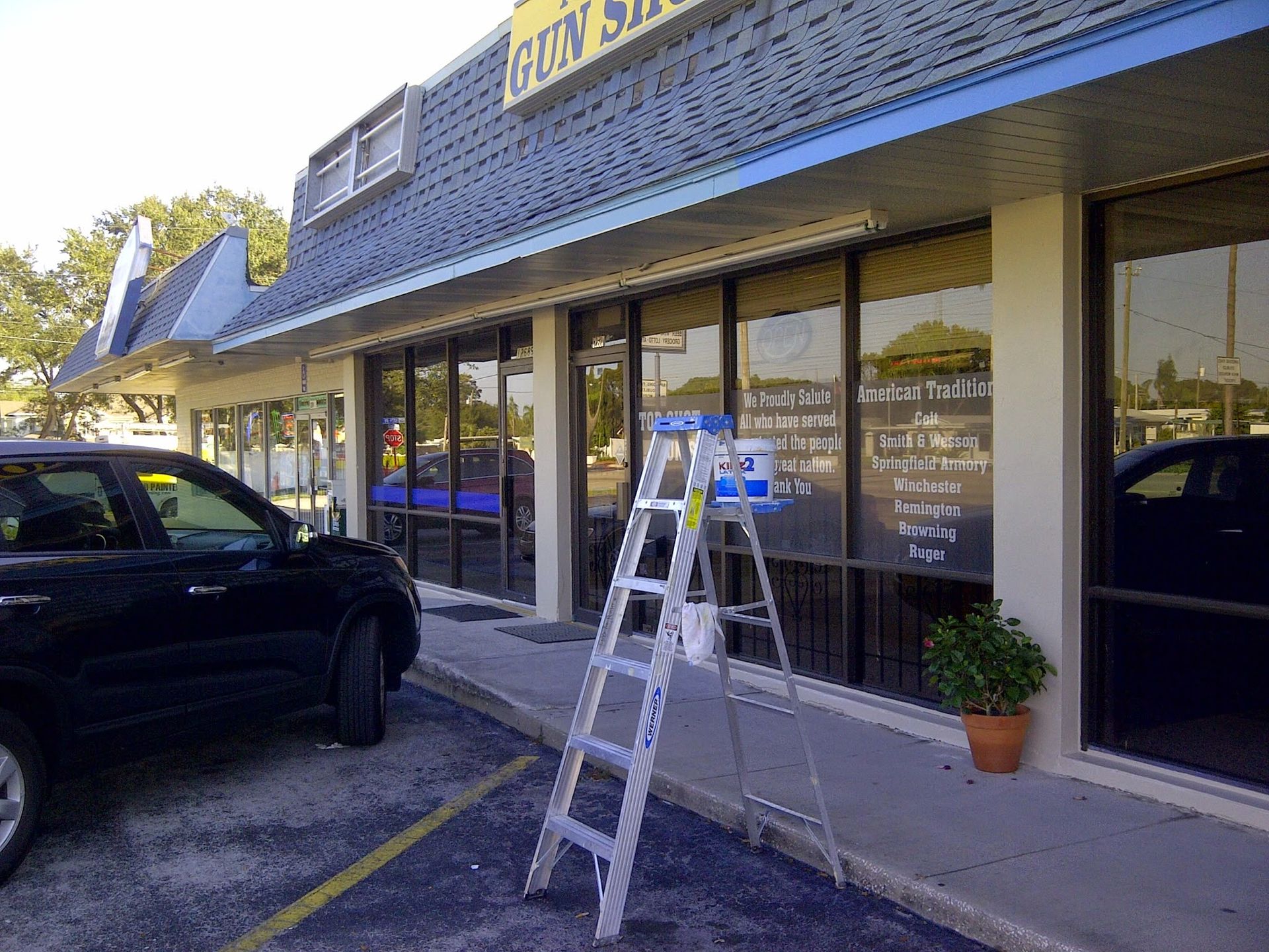 Storefront with a ladder on the sidewalk beside a black SUV and glass windows.