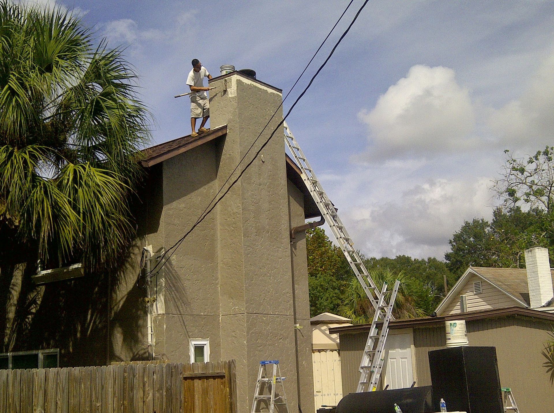 Worker on a ladder spraying water onto the roof of a tan house under a blue sky.