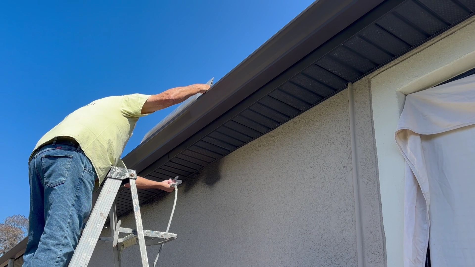 Person on a ladder painting the exterior roof trim of a house under a blue sky