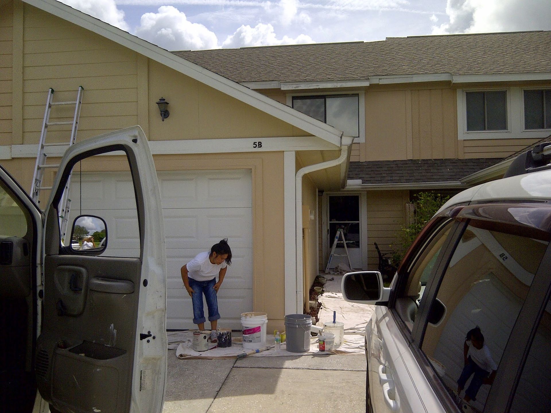 Person painting a garage door outside a suburban house, with paint buckets and vehicles in the driveway.