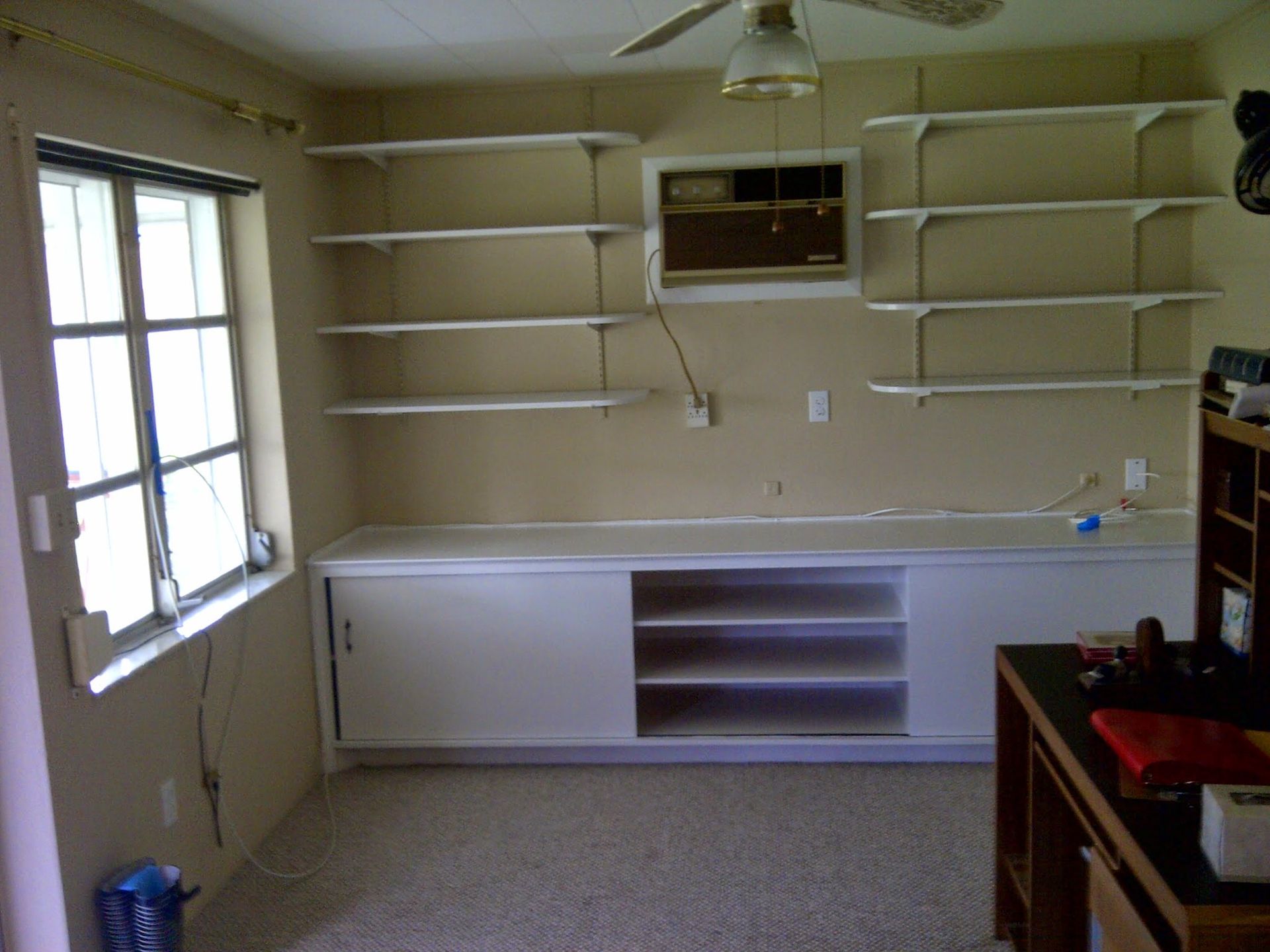 Small room with shelves, a window, and a long white counter under a wall-mounted air conditioner.