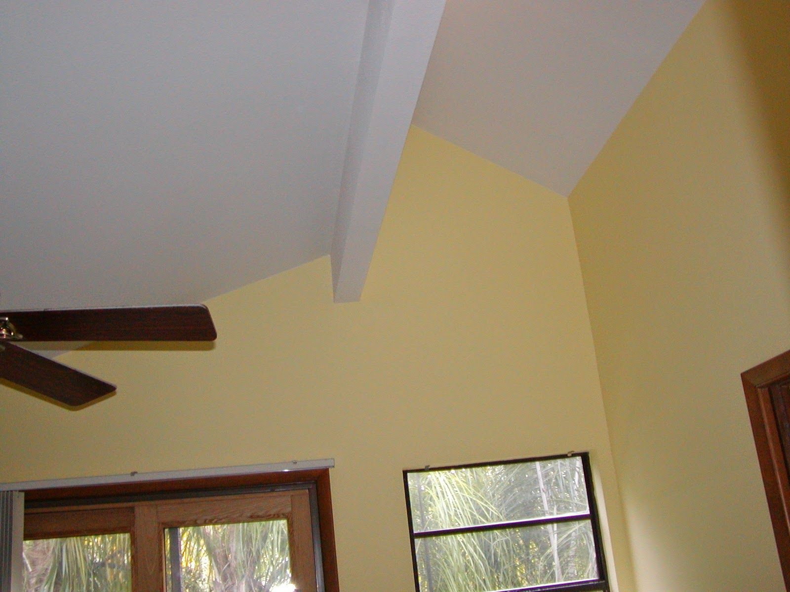 Interior ceiling with sloped white beams, pale yellow walls, a window, and a ceiling fan blade visible